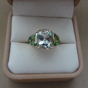 Könnte beinhalten: Ein goldener Ring mit einem großen, hellblauen, rechteckigen Edelstein als Mittelpunkt. Der Ring wird von grünen, blattförmigen Edelsteinen flankiert. Der Ring wird in einer beigen Schmuckschatulle präsentiert.