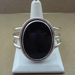 May include: Silver-toned cuff bracelet featuring a large, oval, black stone. The stone is set within a silver frame, and the bracelet has a triple-band design. The bracelet is displayed on a white stand.
