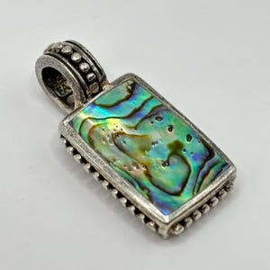 May include: A rectangular pendant with a silver-colored frame and bail. The pendant features a vibrant, iridescent abalone shell inlay with shades of green, blue, and gold. The bail is adorned with small, round details.