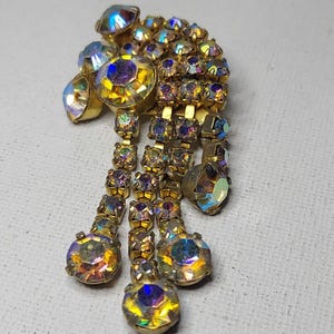 May include: Vintage gold-toned brooch with iridescent rhinestones. The brooch features a cluster of smaller stones and dangling rows of larger, colorful stones. A classic piece of jewelry.