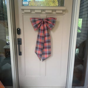 May include: A large decorative bow made of fabric with a plaid pattern in shades of red, grey, and white. The bow is mounted on the center of a light beige front door, which features a black handle, a smart doorbell, and a Ring logo sticker.