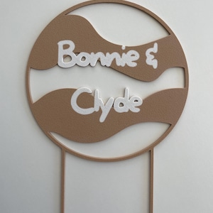 Könnte beinhalten: Ein runder Kuchentopper in Braun und Weiß mit den Namen "Bonnie & Clyde" in weißen Buchstaben. Der Topper hat die Form eines Kreises mit Wellenlinien und ist an zwei dünnen Stäbchen befestigt.