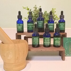 May include: Eight small blue glass bottles with droppers, each containing a different essential oil blend. The bottles are arranged on two wooden shelves, with a mortar and pestle in the foreground. The bottles are labeled with names like "Dream Catcher", "Joyful Balance", and "Brain Waves".
