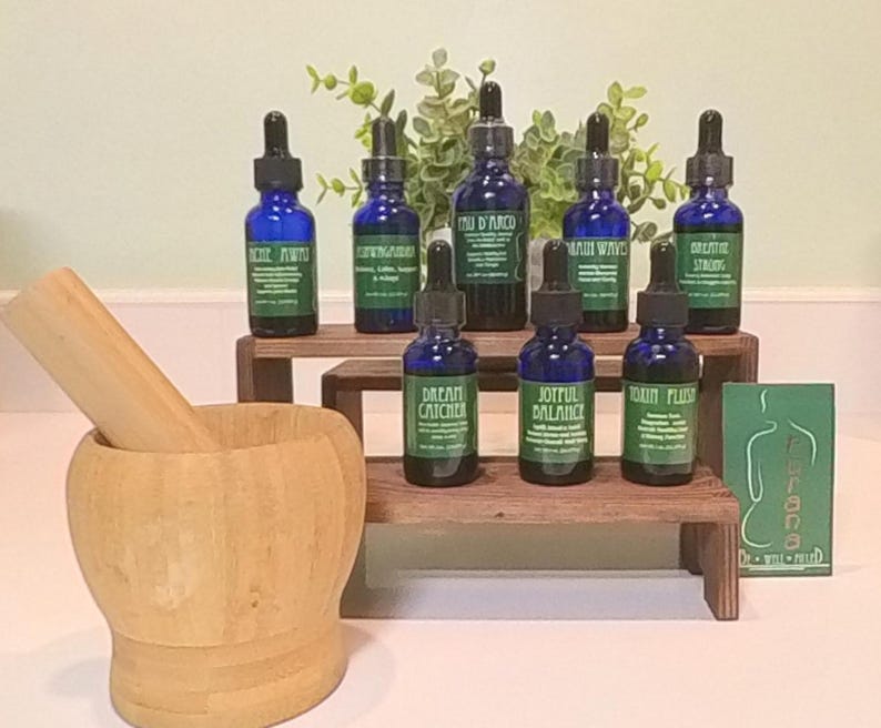 May include: Eight small blue glass bottles with droppers, each containing a different essential oil blend. The bottles are arranged on two wooden shelves, with a mortar and pestle in the foreground. The bottles are labeled with names like "Dream Catcher", "Joyful Balance", and "Brain Waves".
