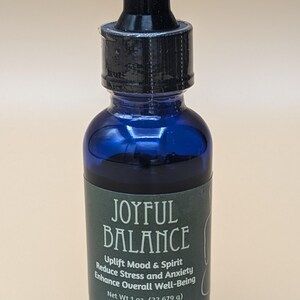 May include: A blue glass bottle with a black dropper top. The bottle has a green label that reads "JOYFUL BALANCE", "Uplift Mood & Spirit", "Reduce Stress and Anxiety", "Enhance Overall Well-Being", and "Net Wt 1 oz. (22.679 g)".
