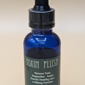 May include: A blue glass bottle with a black dropper top. The label reads "TOXIN FLUSH" and describes the product as a "Remove Toxic Stagnation Assist" that helps with "Overall Healthy Liver & Kidney Function". The bottle contains 1 ounce (22.679 grams) of liquid.