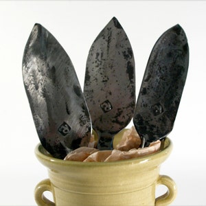 May include: Three black metal leaf-shaped serving utensils with a textured finish are displayed in a light yellow ceramic pot with two handles.