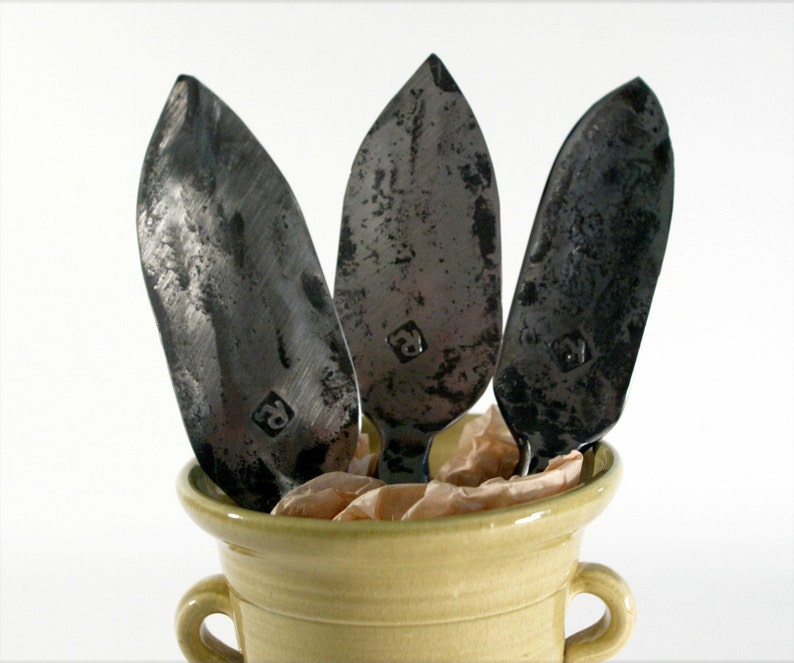 May include: Three black metal leaf-shaped serving utensils with a textured finish are displayed in a light yellow ceramic pot with two handles.