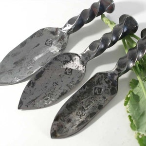 May include: Three hand-forged cheese knives with twisted handles. The knives have a silver finish and are made of metal. Each knife has a small, square mark on the blade.