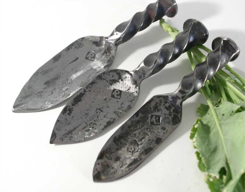 May include: Three hand-forged cheese knives with twisted handles. The knives have a silver finish and are made of metal. Each knife has a small, square mark on the blade.