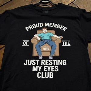 May include: Black t-shirt with the text "PROUD MEMBER OF THE JUST RESTING MY EYES CLUB." The graphic shows a cartoon man in a chair holding a remote and a drink. The name "TONY" is on the chair.
