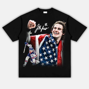 May include: Black t-shirt featuring a graphic of a hockey player wrapped in an American flag. The design includes a signature and a smaller image of the player in action. The shirt is a classic crew neck style.