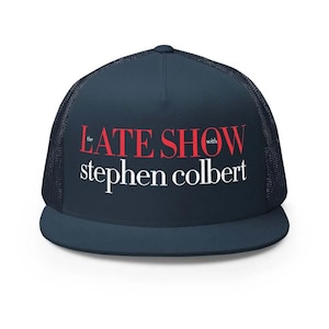 May include: A navy blue trucker hat with a mesh back and a curved brim. The front of the hat features the text "THE LATE SHOW with stephen colbert" in red and white lettering.