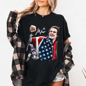 May include: Black t-shirt featuring a graphic of a hockey player holding an American flag. The design includes a signature and the number 86. The player is smiling, and another hockey player is shown in action.