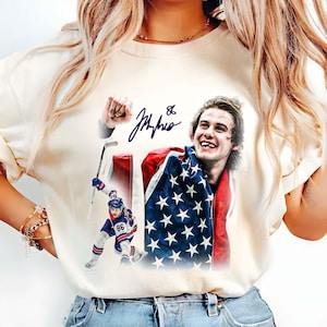 May include: Cream-colored t-shirt featuring a graphic of a hockey player with an American flag. The image includes a signature and the number 86. The player is shown celebrating with a flag draped over his shoulders.