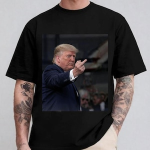 May include: Black t-shirt featuring a photographic print of a man in a suit, raising his middle finger. The image is centered on the shirt, with the man's face and hand in focus. The shirt has short sleeves.