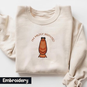 Könnte beinhalten: Cremefarbenes Sweatshirt mit dem gestickten Schriftzug "I'M MILDLY IRRITATED" über einer cartoonartigen, braunen, tropfenförmigen Figur. Das Wort "Embroidery" befindet sich in einem schwarzen Rechteck am unteren Rand.