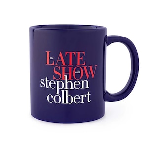 May include: A navy blue ceramic mug with a large handle. The mug features the text "the LATE SHOW with stephen colbert" in red and white lettering.