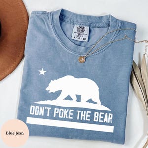 May include: A blue jean colored t-shirt with a white graphic of a bear and the text "DON'T POKE THE BEAR". The shirt has a crew neck and short sleeves. A gold necklace with a pendant is draped across the shirt.
