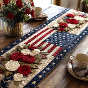 May include: A patriotic table runner featuring the American flag design with red and white stripes and a blue field of stars. The runner is adorned with appliqued red and white roses and green leaves, creating a festive and decorative centerpiece.