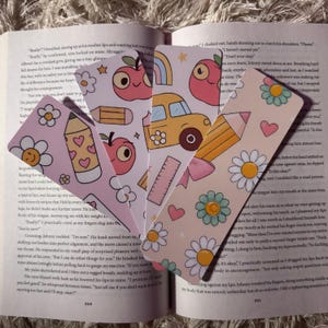 May include: Four colorful bookmarks with school-themed designs, including a yellow school bus, a pencil, and apples. The bookmarks are decorated with hearts, daisies, and rainbows.