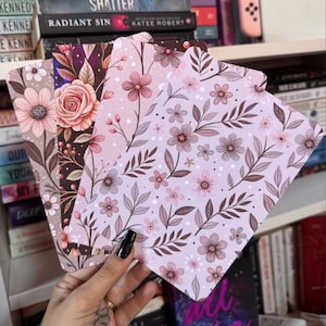 May include: Four floral patterned notebooks in shades of pink, brown, and white are fanned out. The designs include flowers, leaves, and dots. The notebooks are held in a hand with a black painted nail.