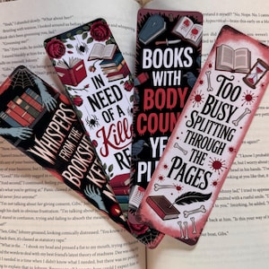 May include: Four decorative bookmarks with gothic-themed designs. One reads "Whispers from the Bookshelf Keeper," another "In Need of a Kill," a third "Books with Body Count," and the last "Too Busy Splitting Through the Pages." The bookmarks are on top of an open book.