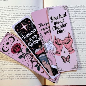 May include: A collection of pink and black bookmarks with various designs and text. One bookmark reads "Romance is my Religion," another says "You had me at Chapter One." The bookmarks feature illustrations of hands, roses, and stars.