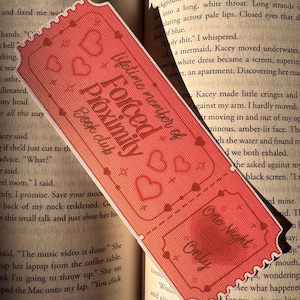 May include: A red bookmark with a ticket design, featuring the text "Forced Proximity Book Club" and "One Night Only." The bookmark has heart and star details, and is placed on an open book.
