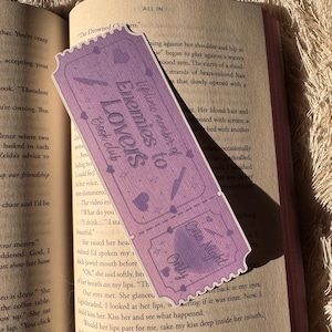 May include: A lavender bookmark shaped like a ticket, featuring the text "Enemies to Lovers Book Club." The design includes hearts, a knife, and the words "One Night Only." The bookmark is resting on the pages of a book.