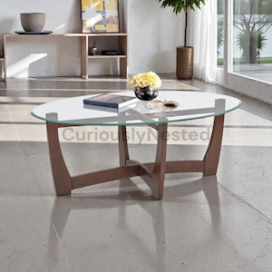 May include: Oval glass-top coffee table with a wooden base. The table has a clear glass top and a dark brown wooden frame. A book and a small vase with yellow flowers are on the table. The text "Curiously Nested" is visible.