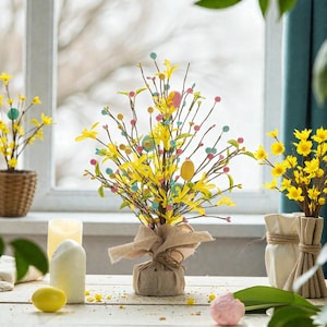 May include: A spring-themed floral arrangement with yellow flowers, colorful pom-poms, and decorative Easter eggs. The arrangement is tied with a burlap ribbon and sits on a white wooden table. Other floral arrangements and candles are also visible.
