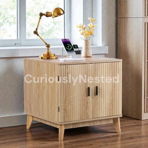May include: A light wood-toned cabinet with a ribbed front design, two black handles, and angled legs. A gold-colored desk lamp, a charging station with a phone and watch, and a vase of yellow flowers sit on top. The text "CuriouslyNested" is visible.