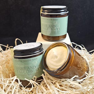 Könnte beinhalten: Drei Gläser mit geschlagenem Talgbalsam mit schwarzen Deckeln und Etiketten mit der Aufschrift "Whipped Tallow Balm" und "Highland Tallow Co." Die Gläser sind bernsteinfarben und mit einem cremigen, cremeweißen Balsam gefüllt. Die Gläser sind auf einem Strohbett angeordnet.
