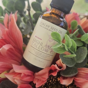 May include: A brown glass bottle of Cacay Seed Oil with a black dropper, labeled "Highland Tallow Co." and "Unscented." The bottle rests among pink and red flowers and green foliage. The label also reads "Face & Neck Retin."