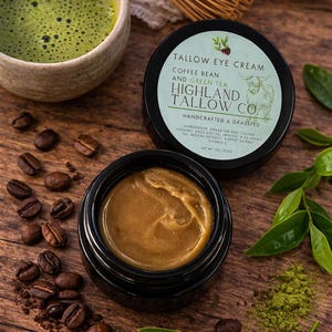 May include: A black-lidded jar of eye cream, labelled "Tallow Eye Cream" with coffee bean and green tea, sits open. Coffee beans, matcha powder, and green leaves are arranged on a wooden surface. The cream inside is golden in colour.