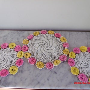 Può includere: Tre centrini bianchi all'uncinetto con un motivo a spirale. Ogni centrino è bordato con fiori all'uncinetto rosa e gialli alternati. I centrini sono esposti su una superficie di marmo.