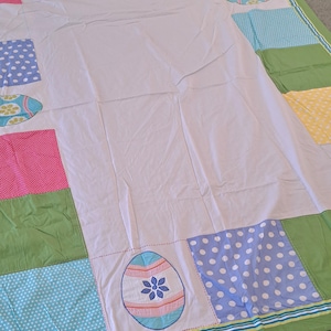 May include: A patchwork quilt with a white center and colorful borders. The quilt features squares of pink, blue, yellow, and green fabric, some with polka dots. Embroidered Easter egg designs and floral accents add detail.