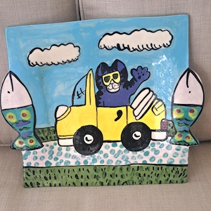 May include: A square ceramic platter featuring a hand-painted scene of a purple cat wearing yellow sunglasses driving a yellow convertible car. The background shows a light blue sky with white clouds, green grass, and two large fish on the sides.