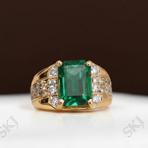 May include: A gold ring with a large, rectangular emerald gemstone at its centre. The ring is embellished with smaller, round, clear gemstones on the sides. It is set on a white surface with a brown backdrop.