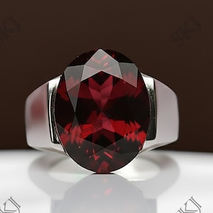 Op de afbeelding: Een zilveren ring met een grote, ovaalvormige, dieprode edelsteen. De edelsteen is het middelpunt, gezet in een eenvoudige, gepolijste zilveren band. De ring wordt getoond op een wit oppervlak met een bruine achtergrond.