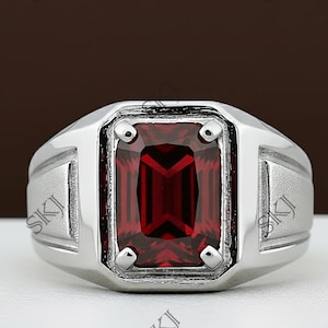 Op de afbeelding: Een zilveren ring met een rechthoekige, dieprode edelsteen in het midden. De ring heeft een brede band en een rechthoekige voorkant, met de edelsteen gezet in een vierkant frame. De ring wordt gepresenteerd op een wit oppervlak.