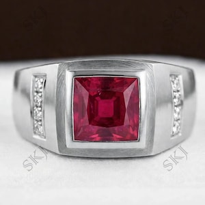 May include: A silver-toned ring featuring a large, square-cut red gemstone set in the centre. The band has a brushed metal finish and includes two vertical rows of small, clear, round-cut accent stones on either side of the central setting.