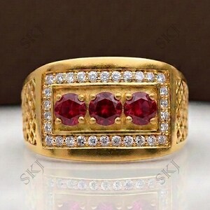 May include: A gold ring featuring a rectangular center panel with three round, deep red rubies arranged horizontally. The central section is framed by two rows of small, clear, sparkling stones. The sides of the ring band have a textured lattice pattern.