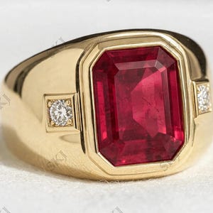 May include: A gold ring featuring a large, rectangular, emerald-cut red gemstone set in the centre. The band includes two small, round, clear accent stones, one on each side of the central setting, embedded within the polished gold metal surface.