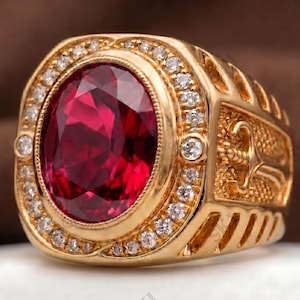 May include: A gold ring featuring a large oval-cut deep red gemstone at the center, surrounded by a halo of small clear round stones. The sides of the gold band are decorated with an embossed sword design and textured detailing throughout the metal.