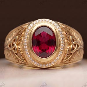 Puede incluir: Un anillo de oro con una gran piedra preciosa de color rojo intenso y corte ovalado en el centro, rodeada por un halo de pequeñas piedras blancas. La banda presenta grabados detallados de plumas y un nudo triquetra a cada lado del engaste.