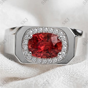 Op de afbeelding: Een zilverkleurige ring met een grote, ovaal geslepen, dieprode edelsteen in het midden. De rode steen wordt vastgehouden door vier pootjes en omringd door een halo van kleine, heldere, rond geslepen steentjes in een rechthoekige zetting.