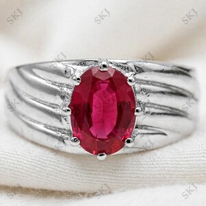 Puede incluir: Un anillo de tono plateado con una piedra preciosa de color rojo intenso de corte ovalado, sujeta por seis garras. La banda tiene un acabado metálico pulido con una serie de ranuras o estrías diagonales que parten del engaste central.