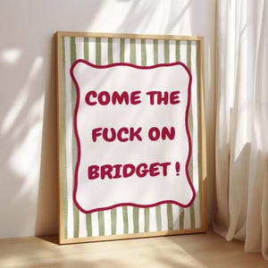 Pode incluir: Impressão artística emoldurada com o texto "COME THE FUCK ON BRIDGET!" em vermelho, sobre um fundo branco com uma borda vermelha. O fundo apresenta listras verticais verdes e brancas, tudo dentro de uma moldura de madeira.